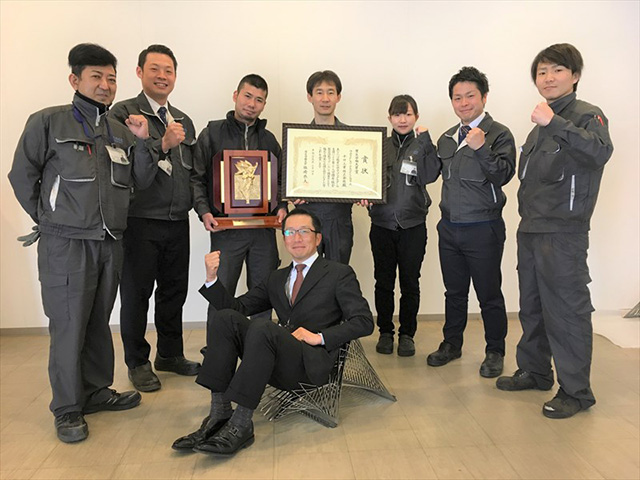 「FLAT-PANEL CHAIR」に座る長澤社長と制作チームの社員との記念写真